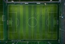Jogadores disputam uma partida de futebol vistos de cima, sobre o campo com suas sombras projetadas.