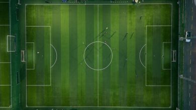 Jogadores disputam uma partida de futebol vistos de cima, sobre o campo com suas sombras projetadas.