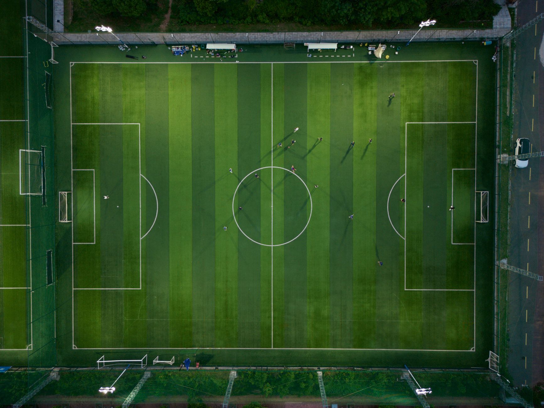Jogadores disputam uma partida de futebol vistos de cima, sobre o campo com suas sombras projetadas.