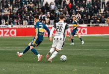 Jogadores disputam uma partida de futebol diante de um estádio cheio.