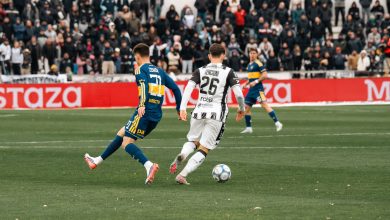 Jogadores disputam uma partida de futebol diante de um estádio cheio.