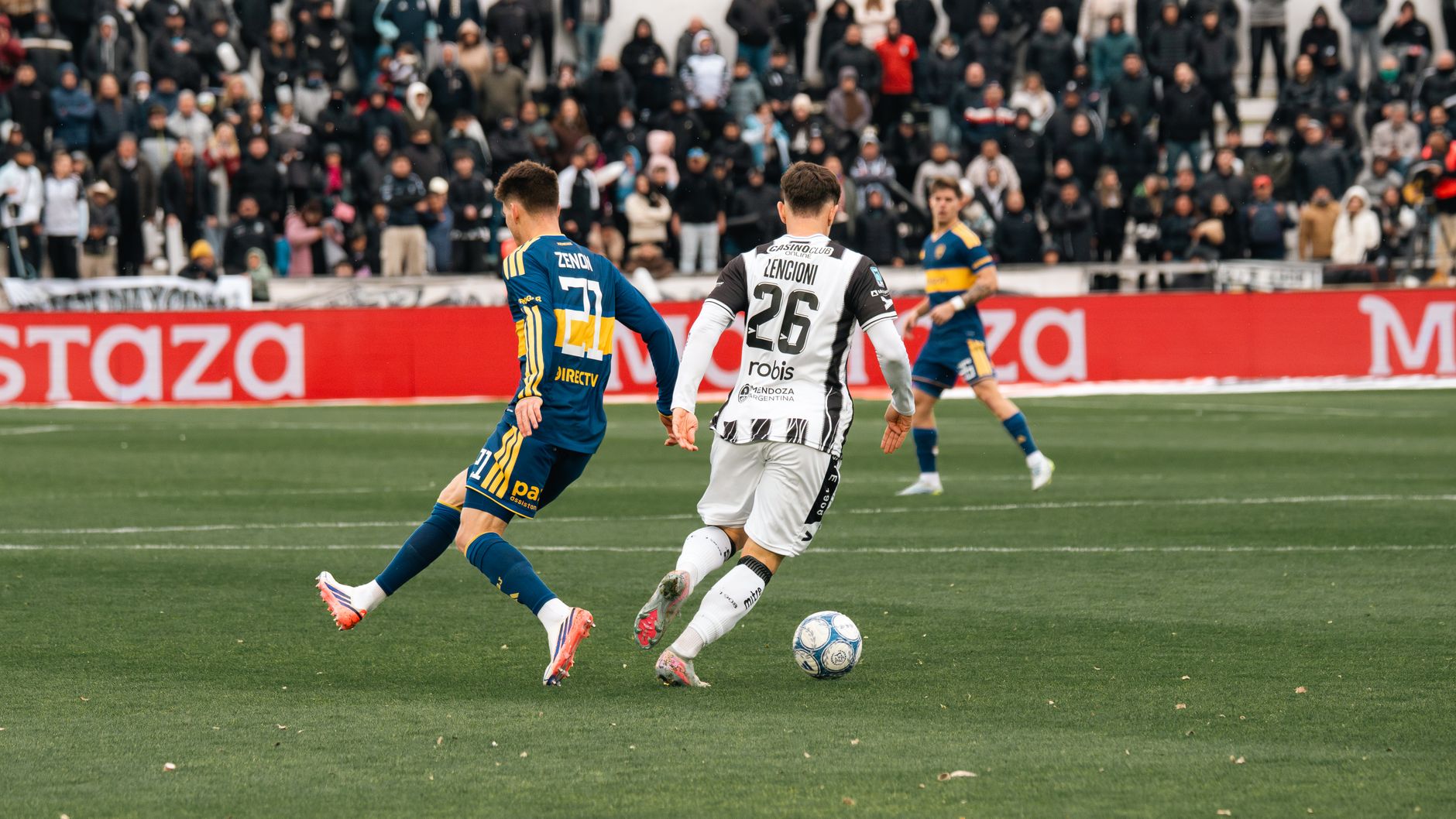Jogadores disputam uma partida de futebol diante de um estádio cheio.